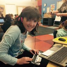 a girl in a classroom wearing headphone at a laptop using a braille display, She is smiling.